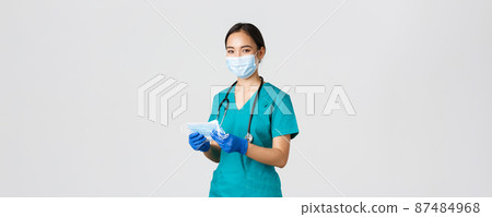 Covid-19, coronavirus disease, healthcare workers concept. Smiling friendly female nurse, physician in scrubs and rubber gloves provide patients with medical masks, white background 87484968