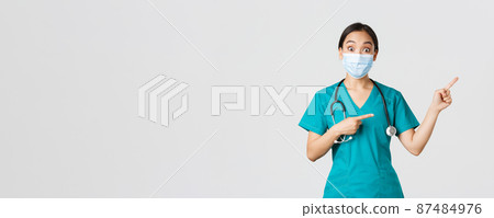 Covid-19, coronavirus disease, healthcare workers concept. Enthusiastic happy asian female doctor, physician or nurse in medical mask and scrubs, pointing upper right corner, show awesome promo 87484976