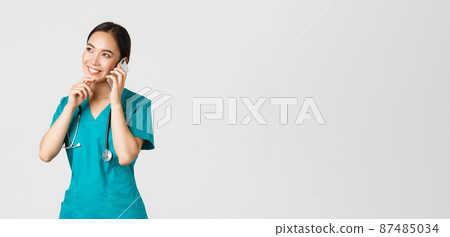 Covid-19, healthcare workers and preventing virus concept. Portrait of smiling asian female doctor, intern in scrubs talking on phone and looking thoughtful, thinking or making choice Covid-19, healthcare workers and preventing virus concept. Portrait of smiling asian female doctor, intern in scrubs talking on phone and looking thoughtful, thinking or making choice 87485034