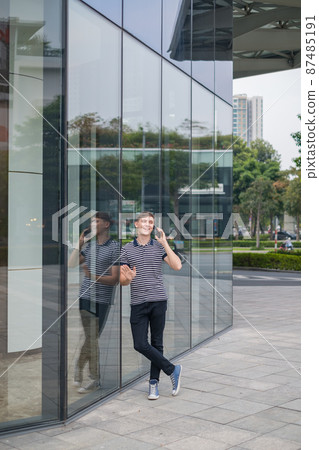 Portrait of young caucasian man in casual clothes standing outdoors and making phone call. Using smartphone, talking by the phone. Urban lifestyle. 87485191