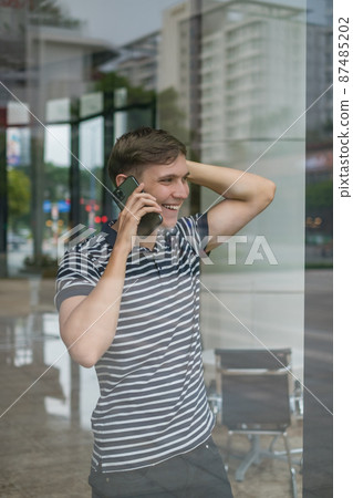 Portrait of young caucasian man in casual clothes making phone call. Photo through the glass. Using smartphone, talking by the phone. Urban lifestyle. 87485202