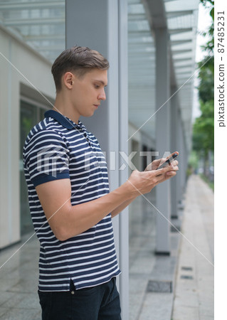 Portrait of Young caucasian guy in casual clothing standing outdoors and looking at his phone. Holding smartphone in his hand. Using phone. 87485231