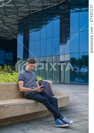 Young caucasian man in casual clothing sitting near modern office building and looking at his phone. High quality photo 87485237