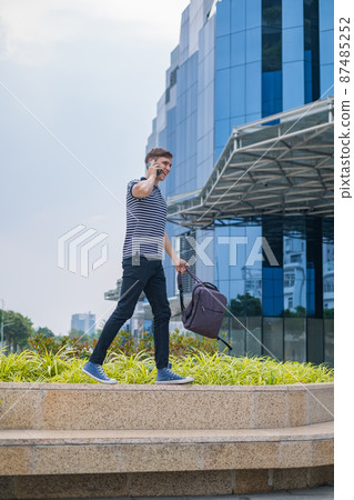 Young caucasian man in casual clothing with the backpack walking near modern office buildings and making phone call. people using phone. High quality Young caucasian man in casual clothing with the backpack walking near modern office buildings and making phone call. people using phone. High quality 87485252