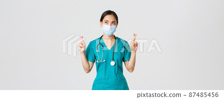 Covid-19, coronavirus disease, healthcare workers concept. Hopeful serious-looking asian doctor, female physician in medical mask and scrubs cross fingers, looking upper left corner 87485456
