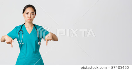 Covid-19, healthcare workers, pandemic concept. Upset and disappointed pouting asian female therapist, nurse in scrubs pointing fingers down, sulking sad, showing something with regret 87485460