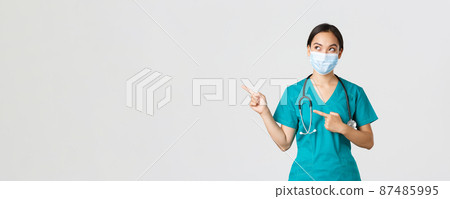 Covid-19, coronavirus disease, healthcare workers concept. Curious pleased asian female doctor, physician in medical mask and gloves, looking and pointing upper left corner with intrigued smile 87485995
