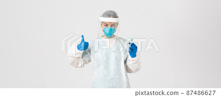 Covid-19, coronavirus disease, healthcare workers concept. Confident serious female asian doctor in personal protective equipment, show thumb-up and hold syringe with vaccine, white background 87486627