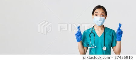 Covid-19, coronavirus disease, healthcare workers concept. Excited and hopeful asian female doctor, nurse in medical mask and rubber gloves cross fingers good luck, makign wish, white background 87486910