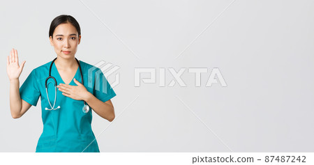 Covid-19, healthcare workers and preventing virus concept. Smiling honest asian female doctor, intern in scrubs giving pledge, raising one arm and hold hand on heart while promise, oath to patient 87487242