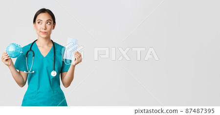 Covid-19, coronavirus disease, healthcare workers concept. Thoughtful smiling asian female doctor, physician in scrubs looking dreamy upper left corner, holding respirator and medical mask 87487395