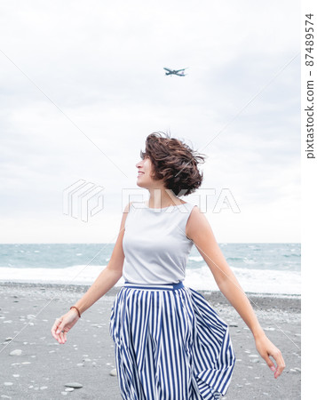 Plane flies in cloudy sky over smiling woman on seaside. Woman with hair ruffled with the wind. Wanderlust concept. Vacation on sea coast. 87489574