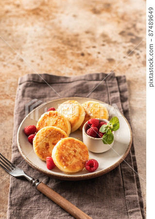 Cottage cheese fritters syrniki with honey topping and raspberry and mint on ceramic plate on napkin on textured background. Text space Cottage cheese fritters syrniki with honey topping and raspberry and mint on ceramic plate on napkin on textured background. Text space 87489649