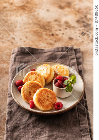 Cottage cheese fritters syrniki with honey topping and raspberry and mint on ceramic plate on napkin on textured background. Text space Cottage cheese fritters syrniki with honey topping and raspberry and mint on ceramic plate on napkin on textured background. Text space 87489650