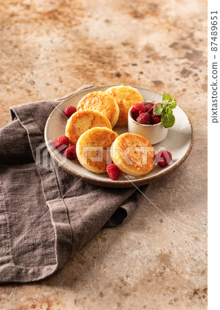 Cottage cheese fritters syrniki with honey topping and raspberry and mint on ceramic plate on napkin on textured background. Text space Cottage cheese fritters syrniki with honey topping and raspberry and mint on ceramic plate on napkin on textured background. Text space 87489651