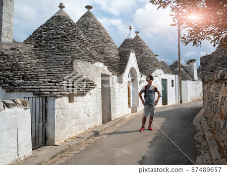 Alberobello, Puglia, Italy. August 2021. Amazing daytime view an alley of the historic village. A Caucasian boy with curly hair and glasses poses for a souvenir photo. Alberobello, Puglia, Italy. August 2021. Amazing daytime view an alley of the historic village. A Caucasian boy with curly hair and glasses poses for a souvenir photo. 87489657