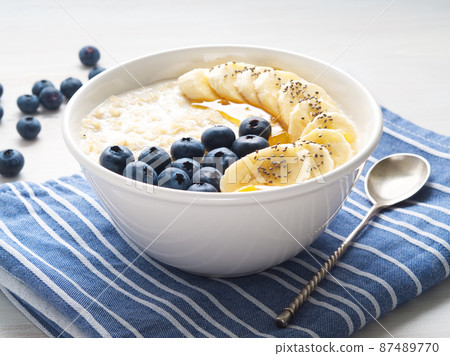 Oatmeal with bananas, blueberries, chia, jam, honey, blue napkin on white wooden background, side view Oatmeal with bananas, blueberries, chia, jam, honey, blue napkin on white wooden background, side view 87489770