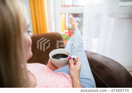 A blonde woman holds a cup of coffee while sitting in a chair. 87489774