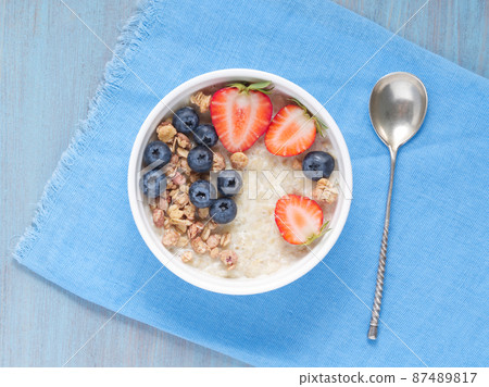 Oatmeal porridge with fresh strawberry, blueberry, granola on blue background. Healthy breakfast. Top view. Oatmeal porridge with fresh strawberry, blueberry, granola on blue background. Healthy breakfast. Top view. 87489817