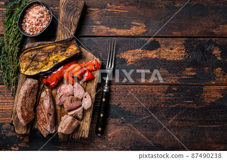 Roasted lamb tenderloin Fillet Meat steak on wooden cutting board. Dark wooden background. Top view. Copy space Roasted lamb tenderloin Fillet Meat steak on wooden cutting board. Dark wooden background. Top view. Copy space 87490238