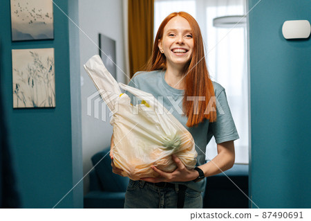 Portrait of happy redhead young woman housewife holding groceries bag and smiling looking at camera, standing on apartment doorstep. Cute female received online order from delivery service. 87490691