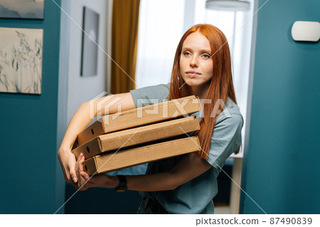 Portrait of pretty young woman holding boxes of hot pizza standing at doorstep to apartment, looking forward. Cute female customer received online order from courier with prepared meal for lunch Portrait of pretty young woman holding boxes of hot pizza standing at doorstep to apartment, looking forward. Cute female customer received online order from courier with prepared meal for lunch 87490839