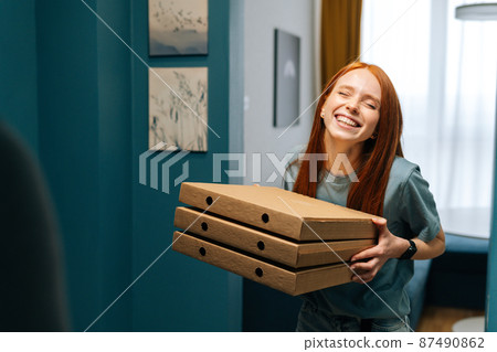 Portrait of excited cheerful young woman holding boxes of hot pizza standing at entrance to apartment. Happy female customer received online order from courier with prepared meal for lunch. 87490862