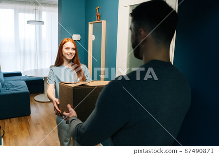 Smiling redhead young woman receiving cardboard box from unrecognizable delivery man on doorstep at apartment. Rear view of courier male delivering parcel package post to female client at home. Smiling redhead young woman receiving cardboard box from unrecognizable delivery man on doorstep at apartment. Rear view of courier male delivering parcel package post to female client at home. 87490871