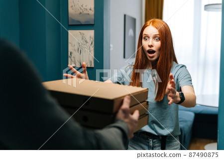 Surprised young woman receiving paper boxes with hot pizza from unrecognizable courier male on doorway at home. Rear view of delivery man delivering boxes with food to female client at apartment. Surprised young woman receiving paper boxes with hot pizza from unrecognizable courier male on doorway at home. Rear view of delivery man delivering boxes with food to female client at apartment. 87490875