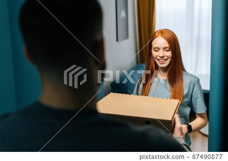 Attractive happy young woman receiving paper boxes with hot pizza from delivery man on doorway at home. Back view of unrecognizable courier male delivering boxes with food to female at apartment. Attractive happy young woman receiving paper boxes with hot pizza from delivery man on doorway at home. Back view of unrecognizable courier male delivering boxes with food to female at apartment. 87490877