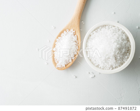 White bowl with large sea salt, wooden spoon, scattered salt crystals on gray stone table. Top view, copy space. 87491872