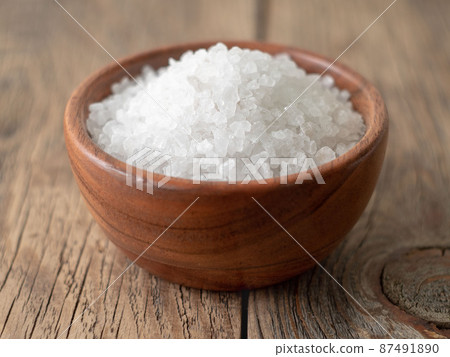 White bowl with large sea salt on brown bright wooden table. Top view, close up, selective focus 87491890