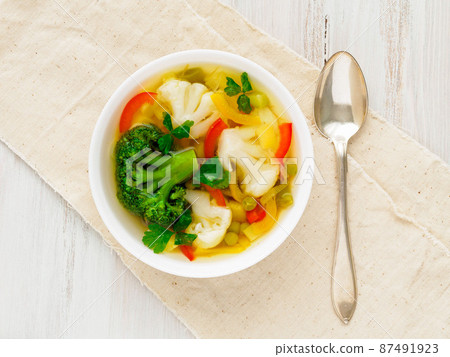 Bright spring vegetable dietary vegetarian soup with cauliflower, broccoli, pepper, carrot, green peas, parsley. Top view, white wood background, linen napkin. Bright spring vegetable dietary vegetarian soup with cauliflower, broccoli, pepper, carrot, green peas, parsley. Top view, white wood background, linen napkin. 87491923
