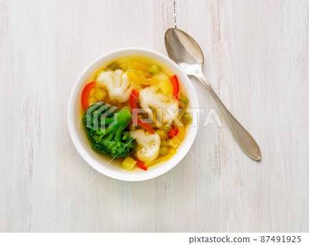 Bright spring vegetable dietary vegetarian soup with cauliflower, broccoli, pepper, carrot, green peas, parsley. Top view, white wood background. Bright spring vegetable dietary vegetarian soup with cauliflower, broccoli, pepper, carrot, green peas, parsley. Top view, white wood background. 87491925