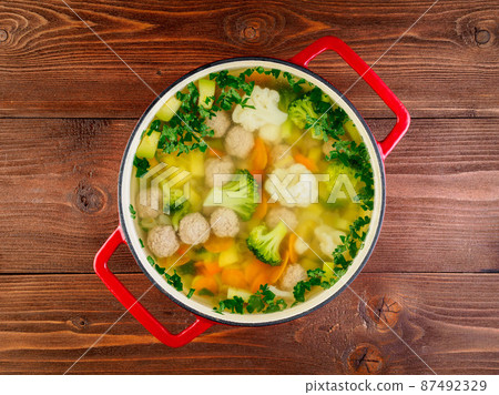 large metal cast red pan with delicious hot soup of vegetables and meatballs, cauliflower, broccoli, carrots, peppers, potatoes, healthy diet food. Brown wooden table, top view. 87492329