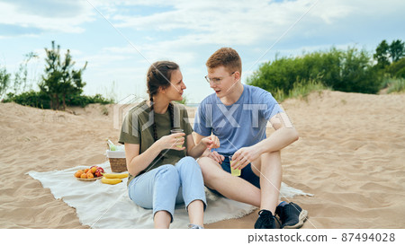 Young adult couple talking and having picnic on beach Young adult couple talking and having picnic on beach 87494028