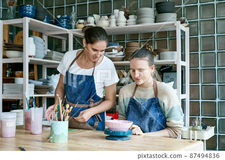 Woman making ceramic pottery. Attractive skilled young lady in apron standing at table and teaching 87494836