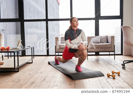 Woman with artificial limb standing at the mat and making lunges while exercising at home 87495194
