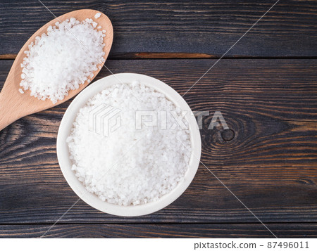 White bowl with large sea salt and wooden spoon on brown dark burnt wooden table. Top view. 87496011