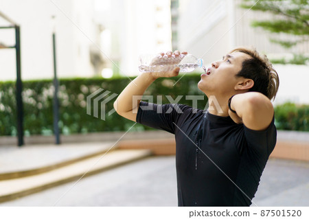 Active Asian sportsman relaxing and drinking a water in bottle after outdoor running or workout. Asian man drink a water during the break from exercise. A healthy man trying hard for cardio workout. 87501520