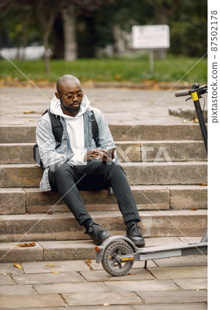 Black man using a phone while sitting on a street with a scooter Black man using a phone while sitting on a street with a scooter 87502178