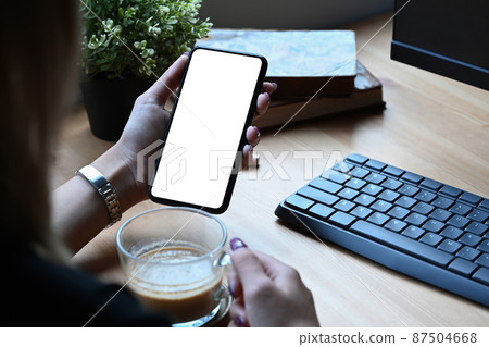 Over shoulder view of young woman drinking coffee and using smart phone at home. Over shoulder view of young woman drinking coffee and using smart phone at home. 87504668