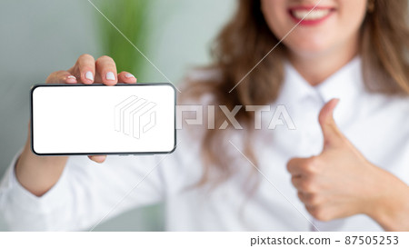 Mobile banking. Office woman. Like gesture. Pretty unrecognizable smiling lady showing smartphone blank screen supportive gesture in light room interior defocused. Mobile banking. Office woman. Like gesture. Pretty unrecognizable smiling lady showing smartphone blank screen supportive gesture in light room interior defocused. 87505253