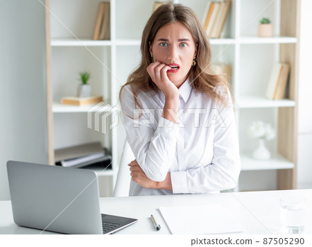Scary emotion. Worried woman. Work problem. Frighten lady in white elegant shirt sitting desk opened laptop biting fingers in light room interior. Scary emotion. Worried woman. Work problem. Frighten lady in white elegant shirt sitting desk opened laptop biting fingers in light room interior. 87505290