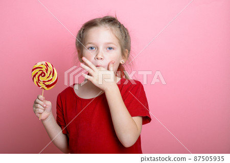 Little cute kid girl lick fingers. Child with lollipop sugar candy. Sticky dirty fingers and girl licks them isolated on pink background. Tasty food Little cute kid girl lick fingers. Child with lollipop sugar candy. Sticky dirty fingers and girl licks them isolated on pink background. Tasty food 87505935