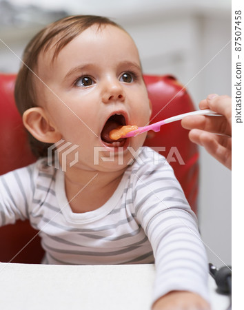 Open wide.... Shot of a cute little baby sitting in a high chair and being fed by her mom. 87507458