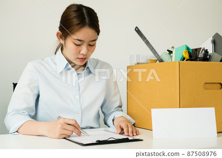 A female office worker, dissatisfied with the termination, packs her belongings in a cardboard box and sits down desperately writing her resignation letter. Concept dismissal and unemployment A female office worker, dissatisfied with the termination, packs her belongings in a cardboard box and sits down desperately writing her resignation letter. Concept dismissal and unemployment 87508976