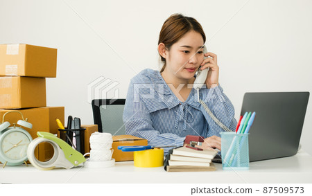 Young Asian woman business owner with many parcel boxes on the table happy online sales job, use the laptop, get an order from customers, take notes, and make arrangements for delivery by post 87509573