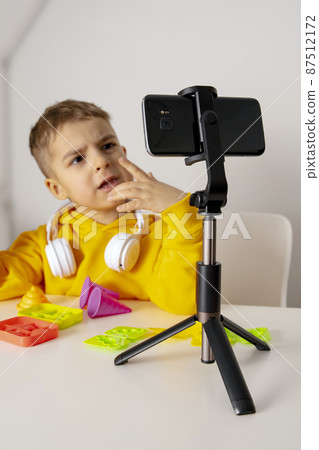 Adorable, cute, little boy blogger recording lifestyle blog, talking to camera of smartphone on tripod. Young influencer filming vlog for his channel. Child makes video for his followers online. Adorable, cute, little boy blogger recording lifestyle blog, talking to camera of smartphone on tripod. Young influencer filming vlog for his channel. Child makes video for his followers online. 87512172