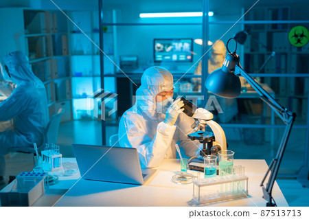 Female scientist in biohazard suit, respirator and gloves using microscope 87513713